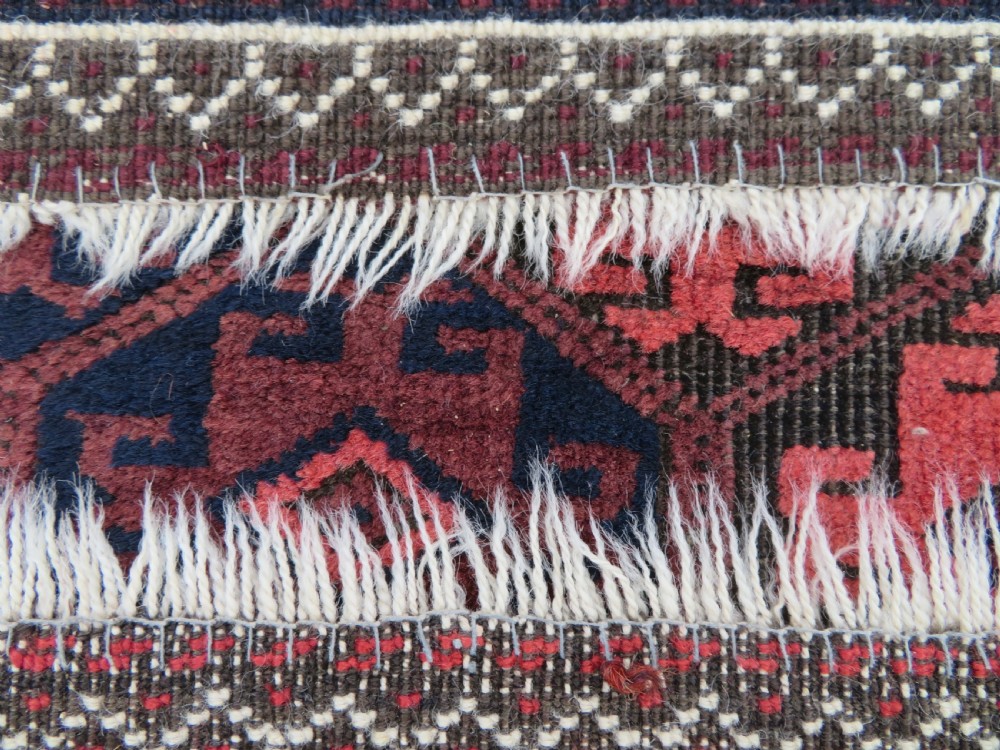 An antique hand-knotted Baluch wool rug featuring repeating geometric diamond motifs in deep red, navy blue, and brown tones with fringed ends. View XV