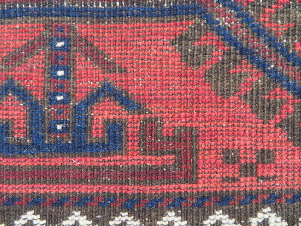 An antique hand-knotted Baluch wool rug featuring repeating geometric diamond motifs in deep red, navy blue, and brown tones with fringed ends. View XVI