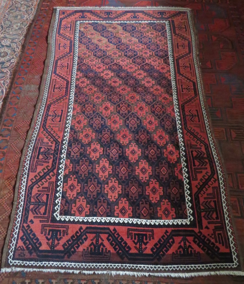 An antique hand-knotted Baluch wool rug featuring repeating geometric diamond motifs in deep red, navy blue, and brown tones with fringed ends. View XVIII