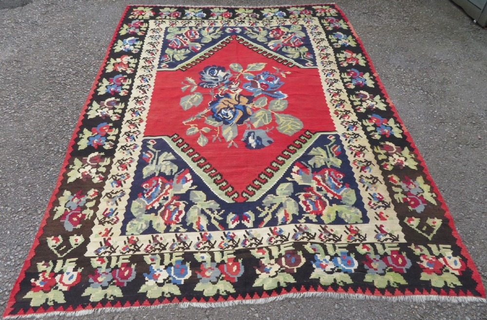An antique flatweave wool rug featuring a central red panel with blue floral motifs, bordered by dark navy and cream botanical patterns.