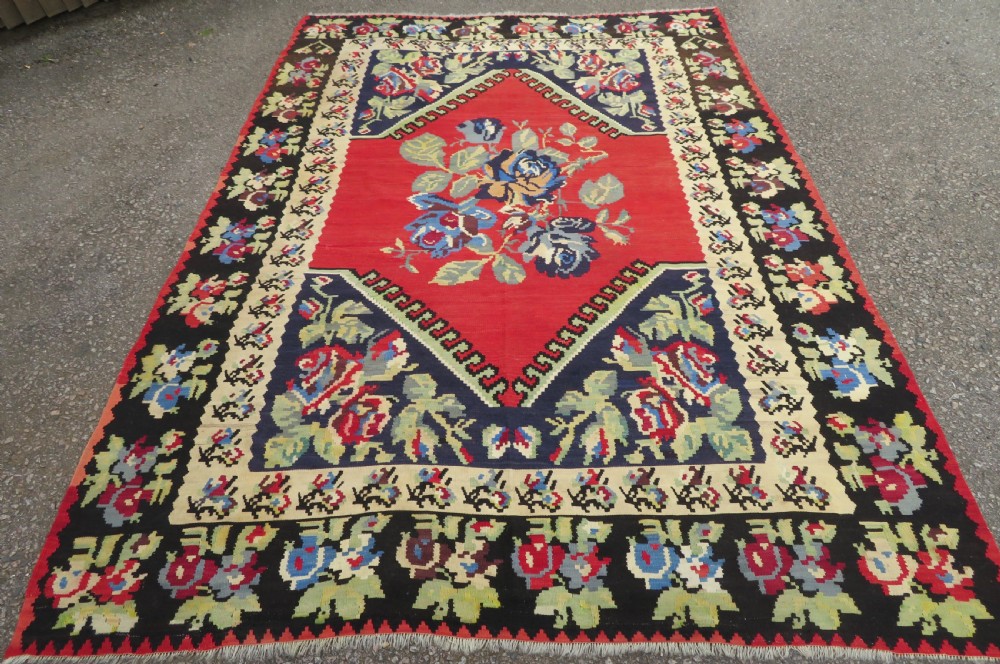 An antique flatweave wool rug featuring a central red panel with blue floral motifs, bordered by dark navy and cream botanical patterns. View II