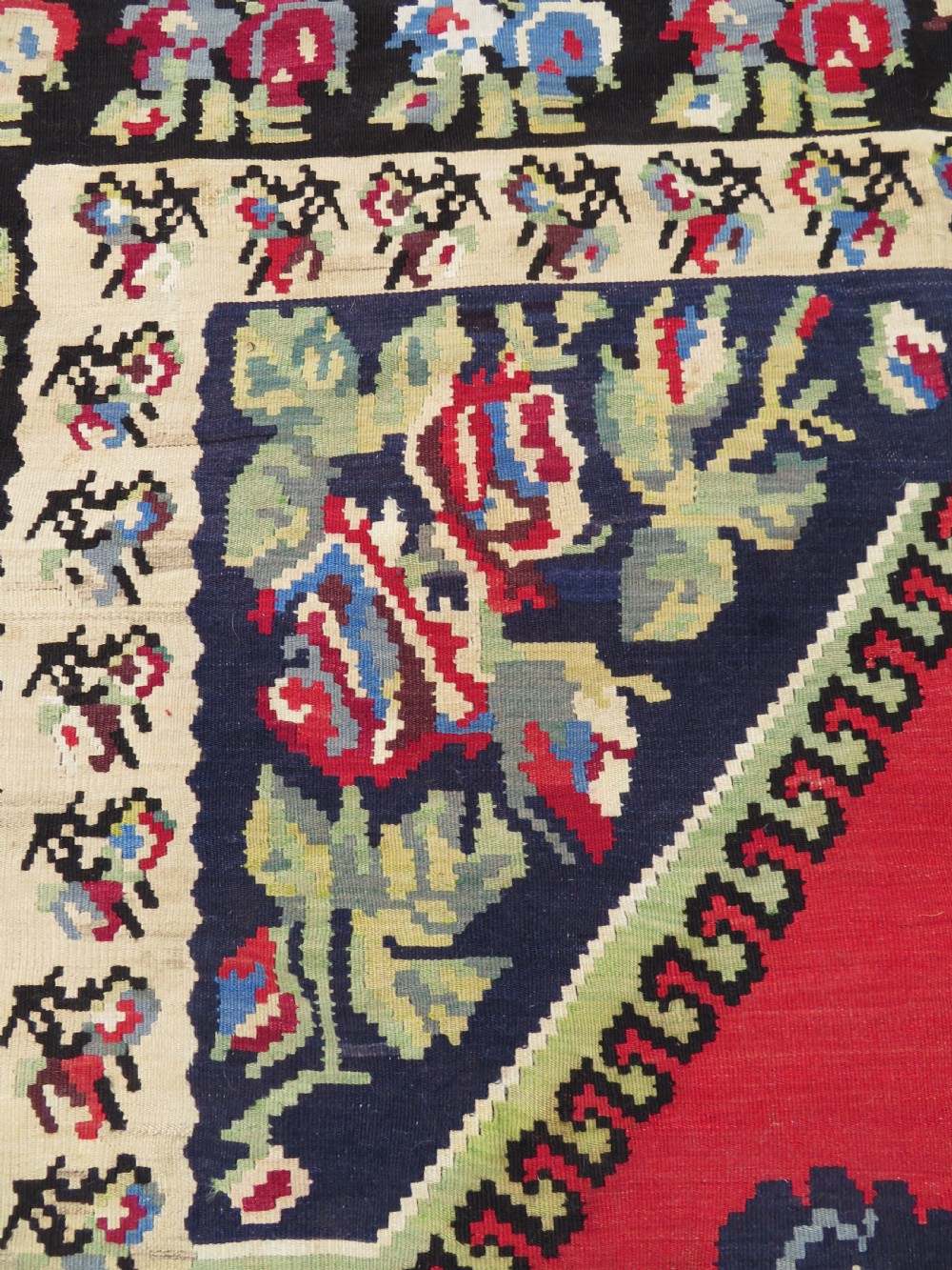 An antique flatweave wool rug featuring a central red panel with blue floral motifs, bordered by dark navy and cream botanical patterns. View IV