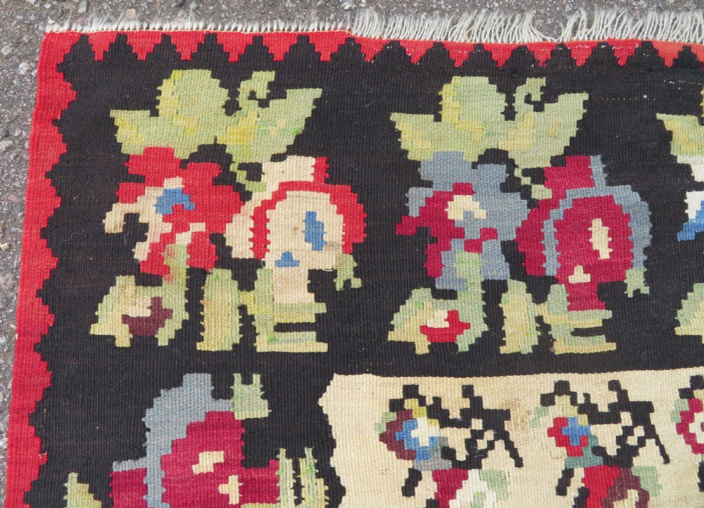An antique flatweave wool rug featuring a central red panel with blue floral motifs, bordered by dark navy and cream botanical patterns. View V