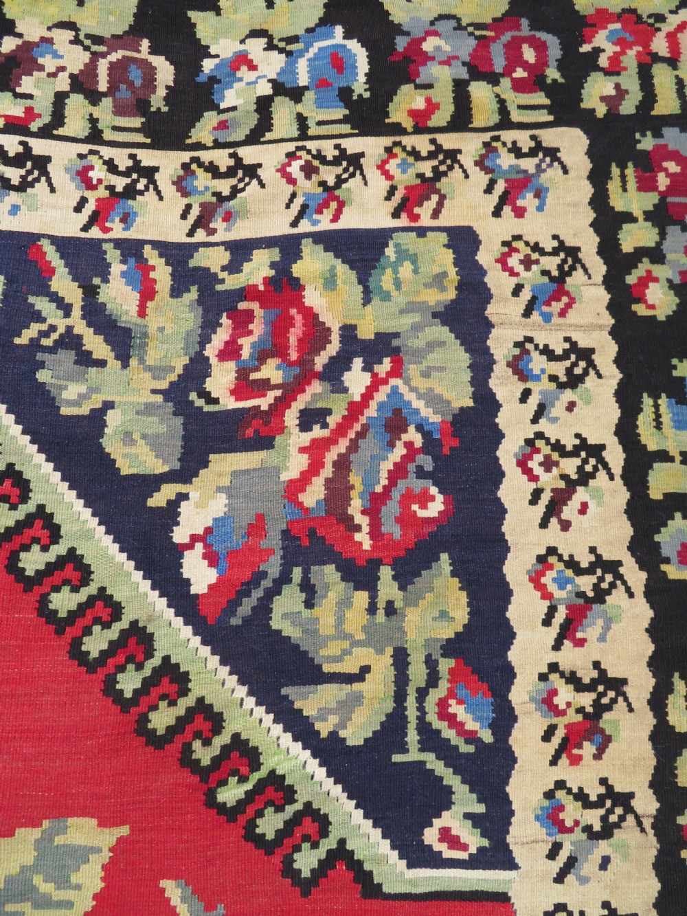 An antique flatweave wool rug featuring a central red panel with blue floral motifs, bordered by dark navy and cream botanical patterns. View VI