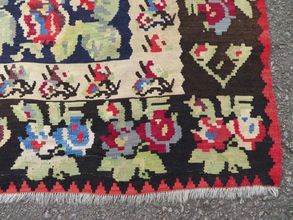 An antique flatweave wool rug featuring a central red panel with blue floral motifs, bordered by dark navy and cream botanical patterns. View IX