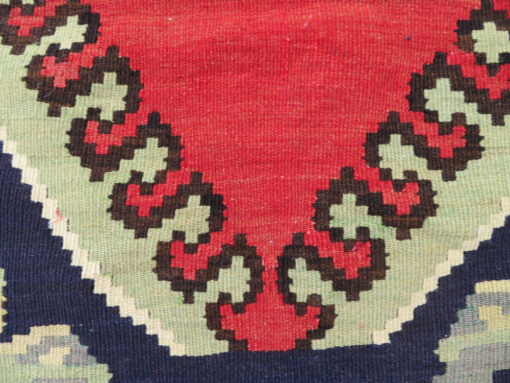 An antique flatweave wool rug featuring a central red panel with blue floral motifs, bordered by dark navy and cream botanical patterns. View XI