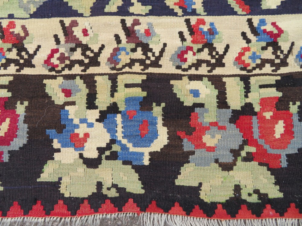An antique flatweave wool rug featuring a central red panel with blue floral motifs, bordered by dark navy and cream botanical patterns. View XII
