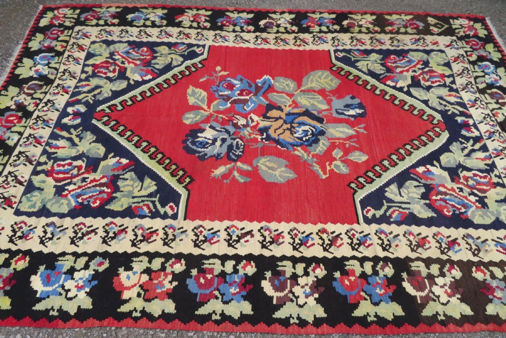 An antique flatweave wool rug featuring a central red panel with blue floral motifs, bordered by dark navy and cream botanical patterns. View XVI
