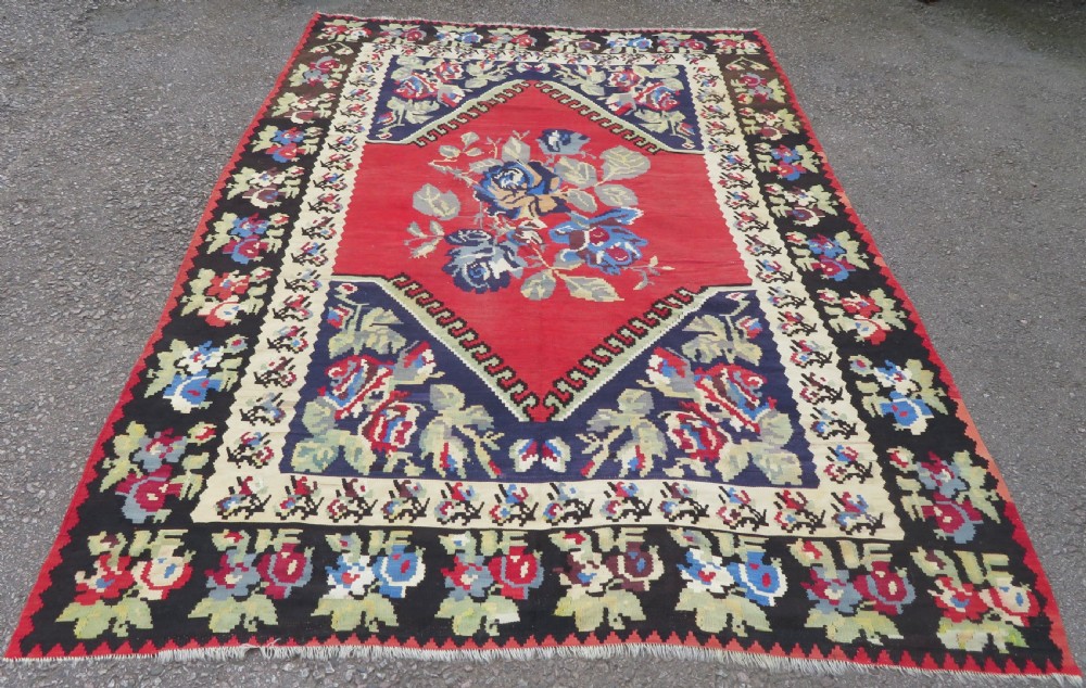An antique flatweave wool rug featuring a central red panel with blue floral motifs, bordered by dark navy and cream botanical patterns. View XVII