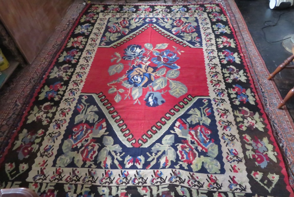 An antique flatweave wool rug featuring a central red panel with blue floral motifs, bordered by dark navy and cream botanical patterns. View XXI