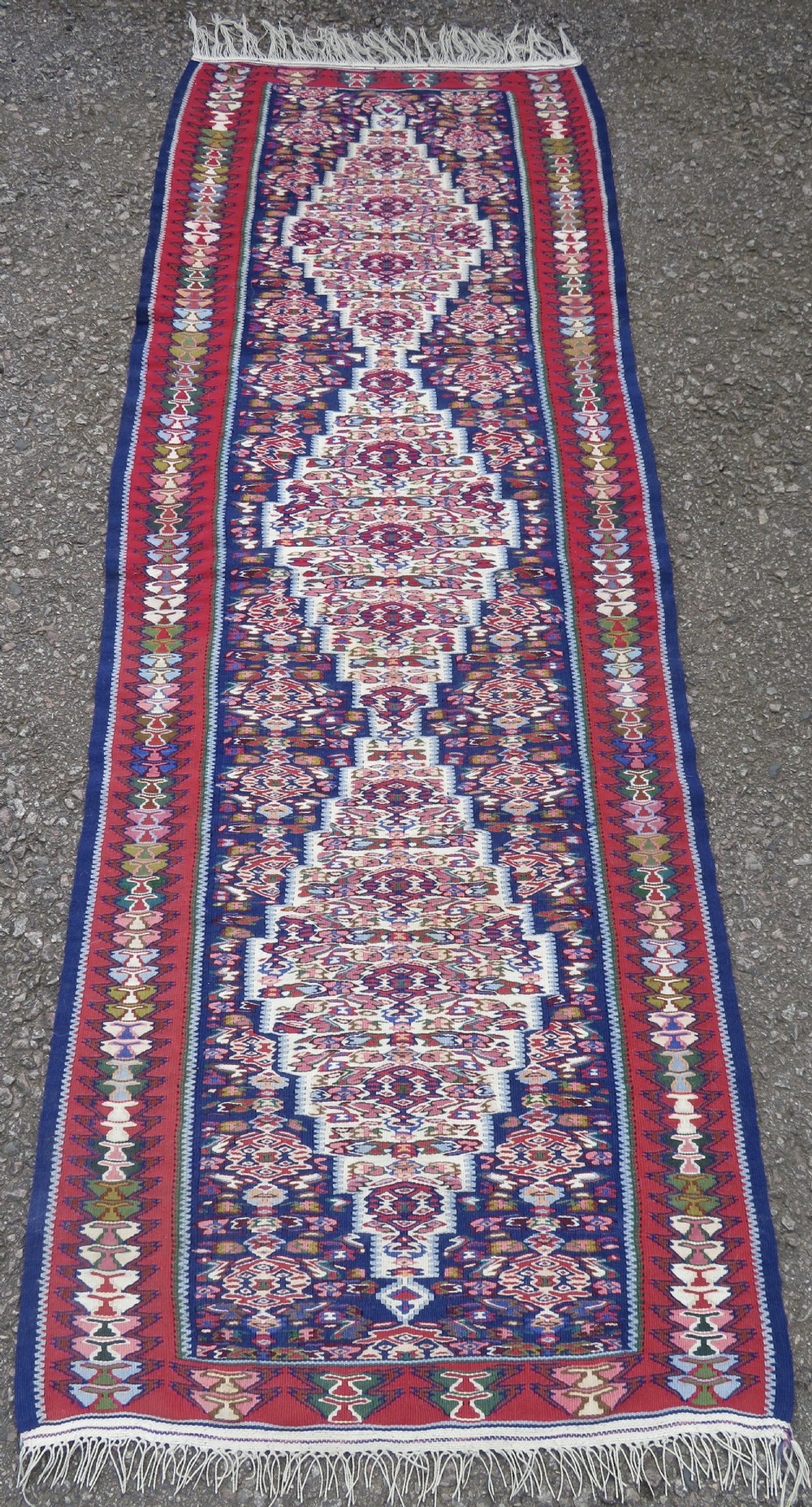 A long, narrow hand-woven Persian Senneh kilim runner featuring a stepped geometric diamond pattern in cream and navy blue bordered by a red outer frame with fringed ends.