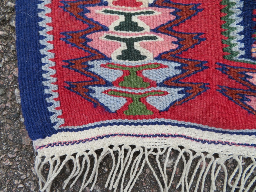 A long, narrow hand-woven Persian Senneh kilim runner featuring a stepped geometric diamond pattern in cream and navy blue bordered by a red outer frame with fringed ends. View IV