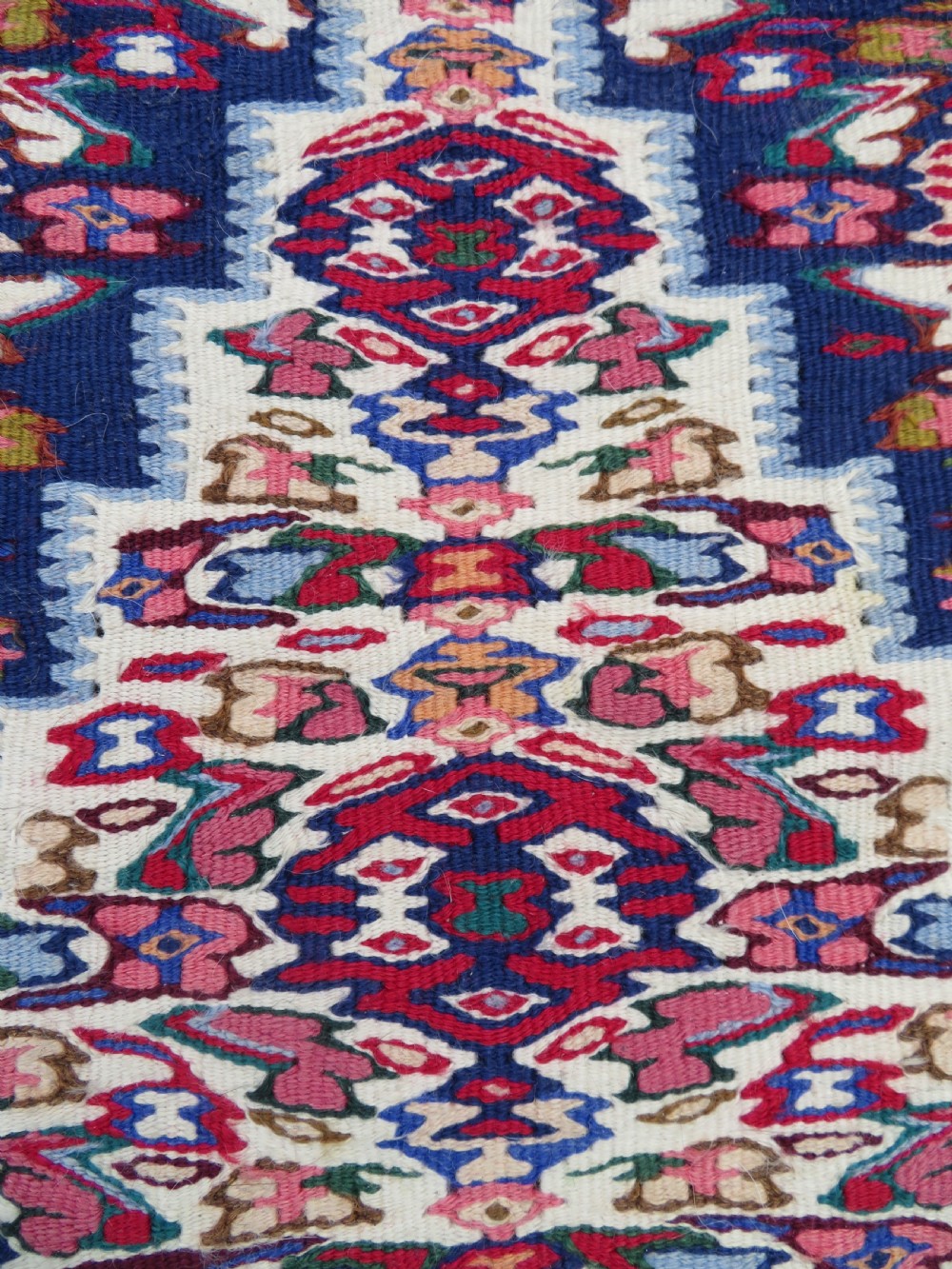 A long, narrow hand-woven Persian Senneh kilim runner featuring a stepped geometric diamond pattern in cream and navy blue bordered by a red outer frame with fringed ends. View V