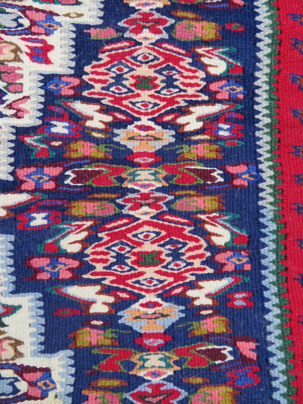 A long, narrow hand-woven Persian Senneh kilim runner featuring a stepped geometric diamond pattern in cream and navy blue bordered by a red outer frame with fringed ends. View VI