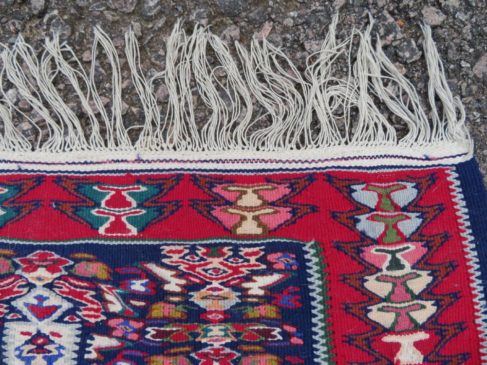 A long, narrow hand-woven Persian Senneh kilim runner featuring a stepped geometric diamond pattern in cream and navy blue bordered by a red outer frame with fringed ends. View VII