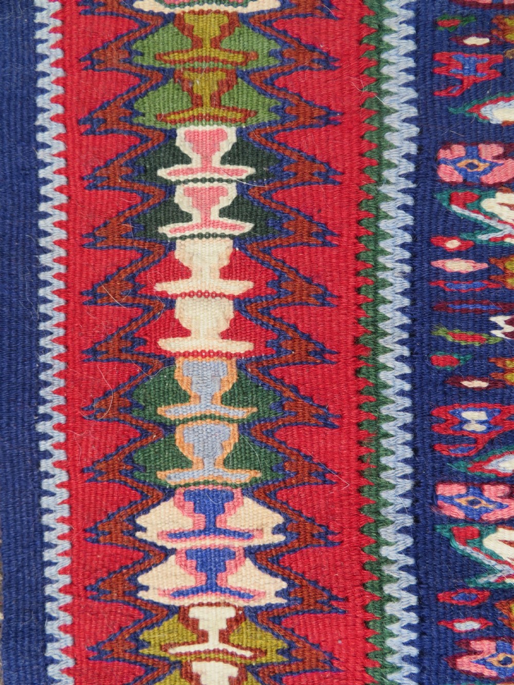 A long, narrow hand-woven Persian Senneh kilim runner featuring a stepped geometric diamond pattern in cream and navy blue bordered by a red outer frame with fringed ends. View IX
