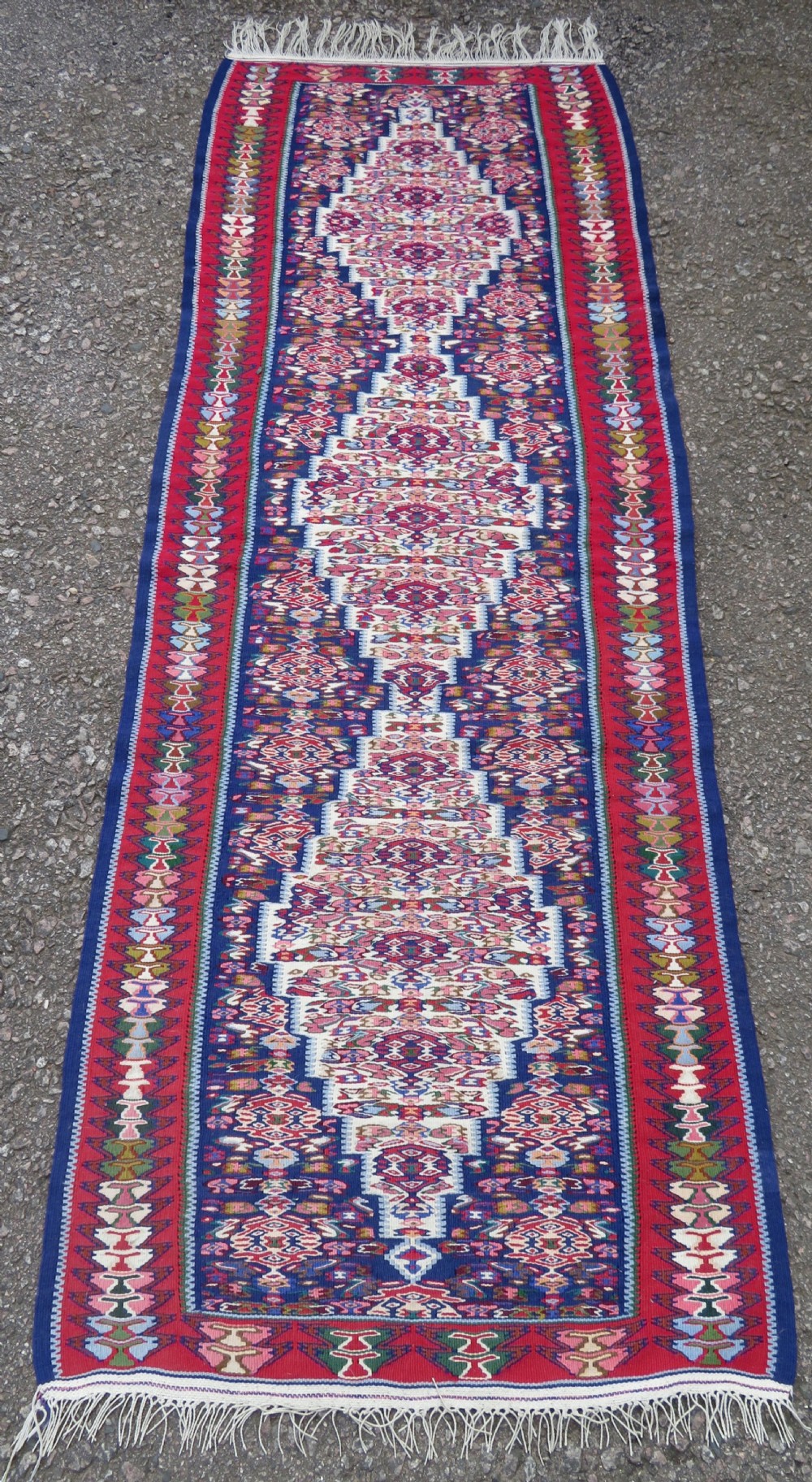 A long, narrow hand-woven Persian Senneh kilim runner featuring a stepped geometric diamond pattern in cream and navy blue bordered by a red outer frame with fringed ends. View X