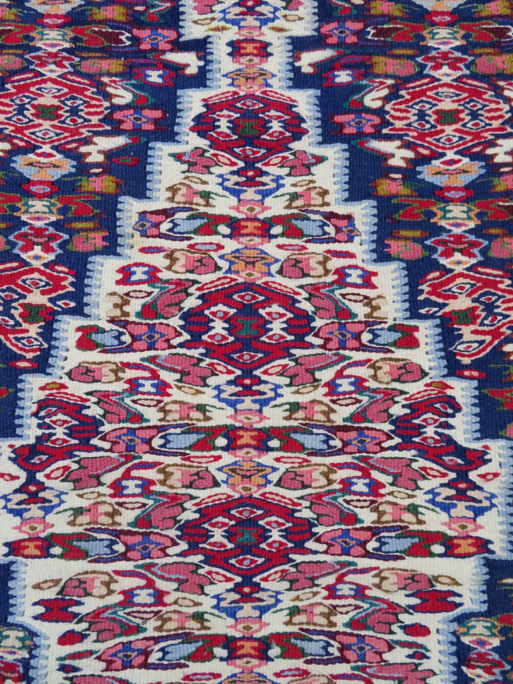 A long, narrow hand-woven Persian Senneh kilim runner featuring a stepped geometric diamond pattern in cream and navy blue bordered by a red outer frame with fringed ends. View XII