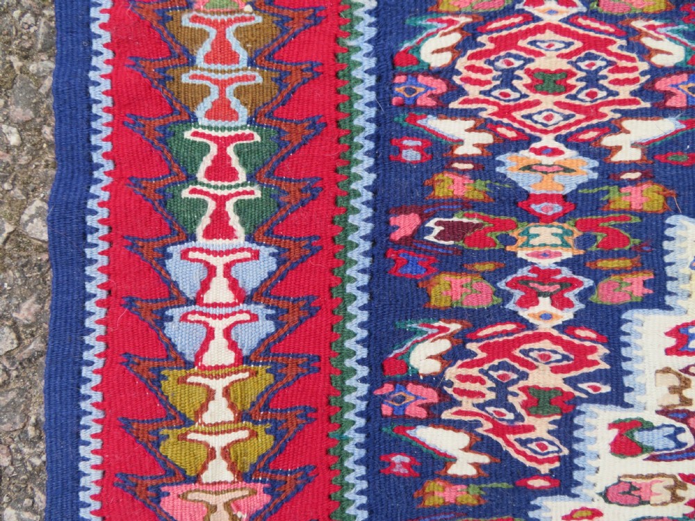 A long, narrow hand-woven Persian Senneh kilim runner featuring a stepped geometric diamond pattern in cream and navy blue bordered by a red outer frame with fringed ends. View XIII