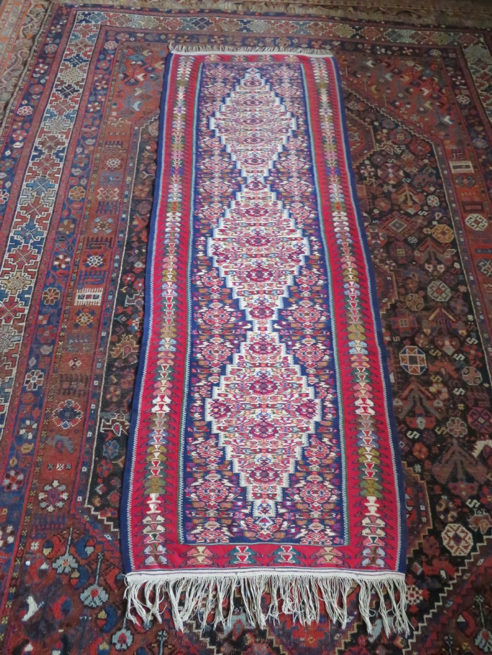 A long, narrow hand-woven Persian Senneh kilim runner featuring a stepped geometric diamond pattern in cream and navy blue bordered by a red outer frame with fringed ends. View XVI