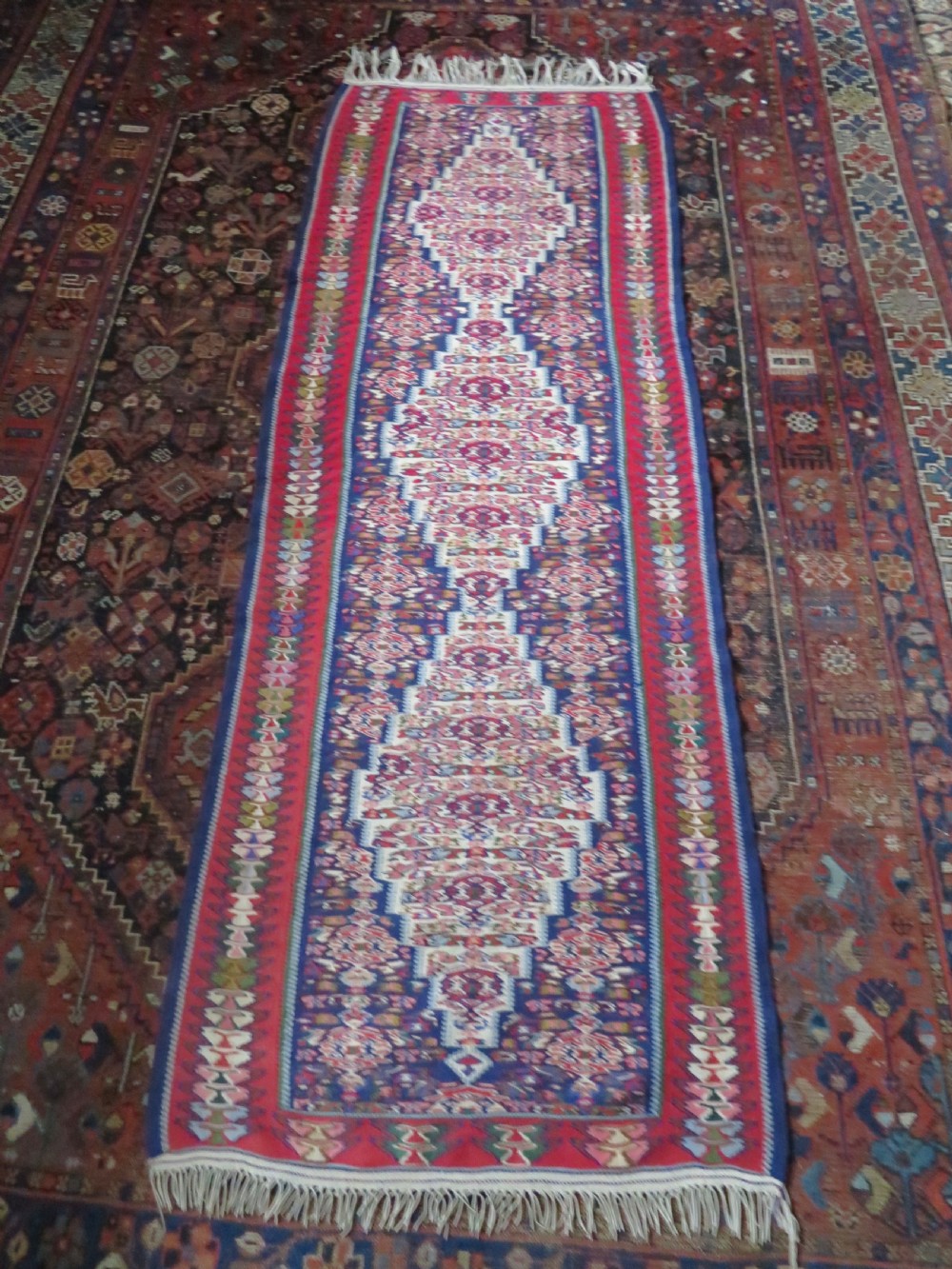 A long, narrow hand-woven Persian Senneh kilim runner featuring a stepped geometric diamond pattern in cream and navy blue bordered by a red outer frame with fringed ends. View XVII