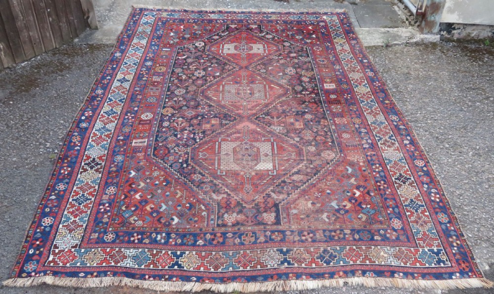 An antique Persian Khamseh wool carpet featuring a dark navy blue field with three stacked diamond medallions and geometric borders in terracotta red and ivory.