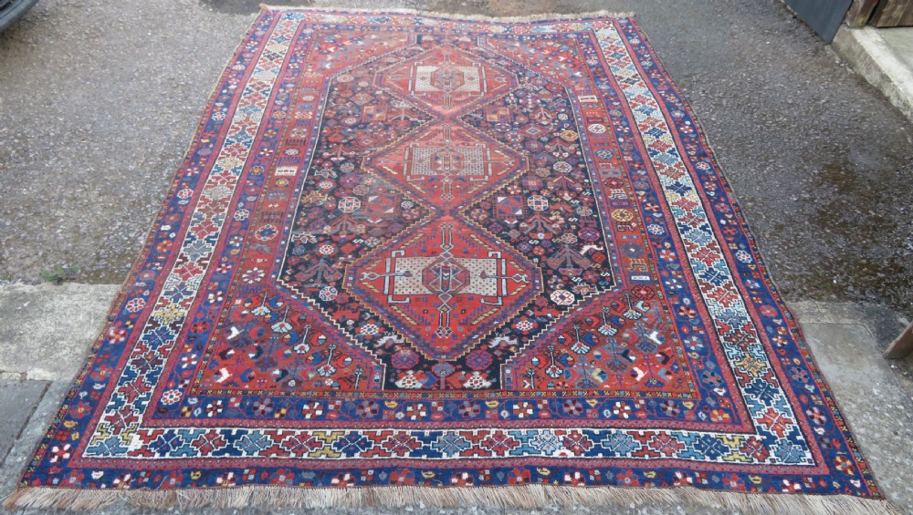 An antique Persian Khamseh wool carpet featuring a dark navy blue field with three stacked diamond medallions and geometric borders in terracotta red and ivory. View II