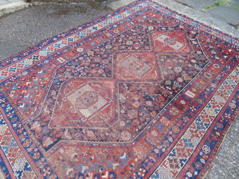 An antique Persian Khamseh wool carpet featuring a dark navy blue field with three stacked diamond medallions and geometric borders in terracotta red and ivory. View IV