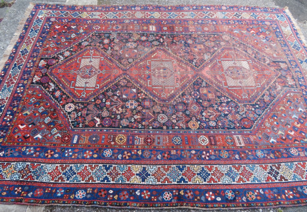An antique Persian Khamseh wool carpet featuring a dark navy blue field with three stacked diamond medallions and geometric borders in terracotta red and ivory. View V