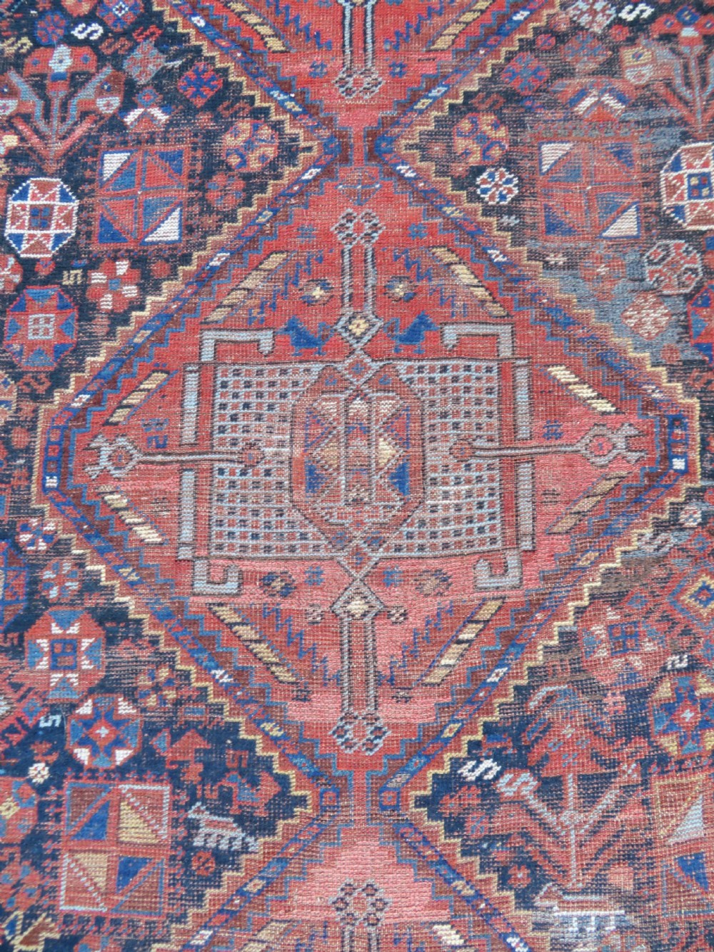 An antique Persian Khamseh wool carpet featuring a dark navy blue field with three stacked diamond medallions and geometric borders in terracotta red and ivory. View VI