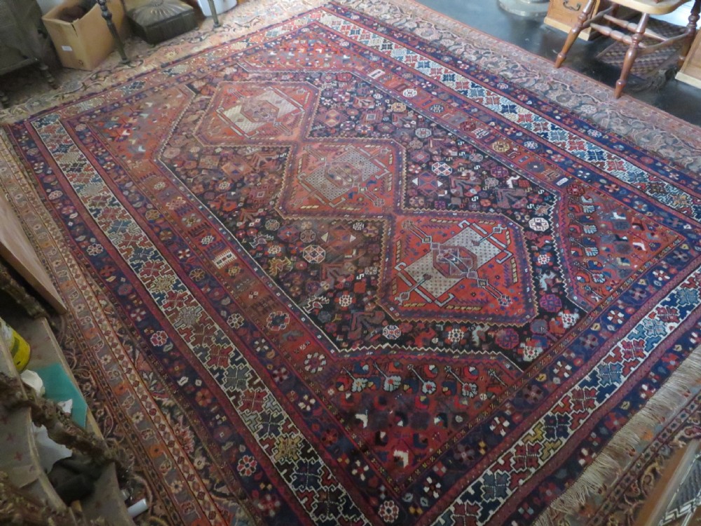 An antique Persian Khamseh wool carpet featuring a dark navy blue field with three stacked diamond medallions and geometric borders in terracotta red and ivory. View XXX