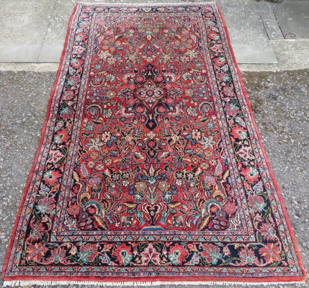 An antique hand-knotted Persian Sarouk wool rug featuring a red field with a central navy medallion and dense floral motifs within a dark border.