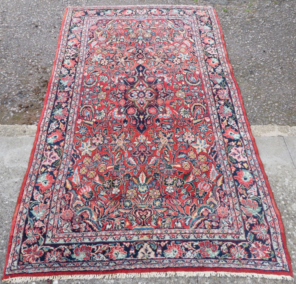 An antique hand-knotted Persian Sarouk wool rug featuring a red field with a central navy medallion and dense floral motifs within a dark border. View II