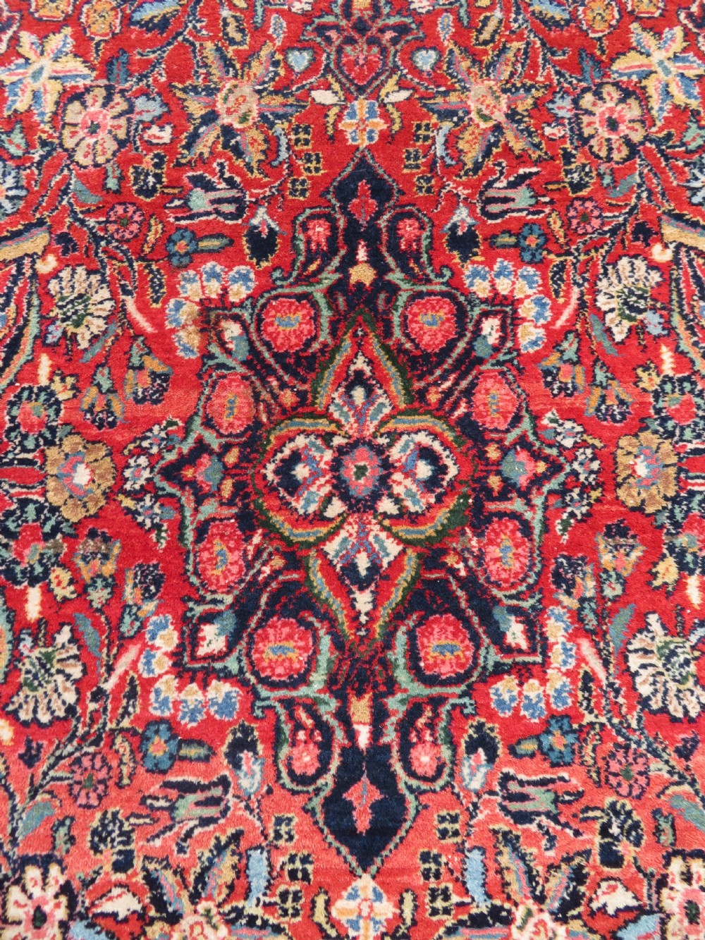 An antique hand-knotted Persian Sarouk wool rug featuring a red field with a central navy medallion and dense floral motifs within a dark border. View III