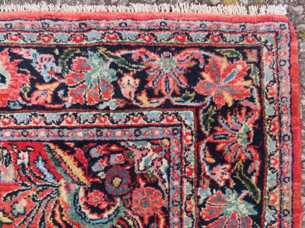 An antique hand-knotted Persian Sarouk wool rug featuring a red field with a central navy medallion and dense floral motifs within a dark border. View IV