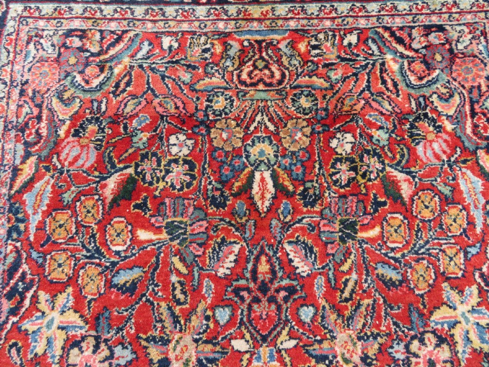 An antique hand-knotted Persian Sarouk wool rug featuring a red field with a central navy medallion and dense floral motifs within a dark border. View VI