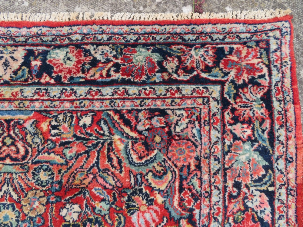 An antique hand-knotted Persian Sarouk wool rug featuring a red field with a central navy medallion and dense floral motifs within a dark border. View VII