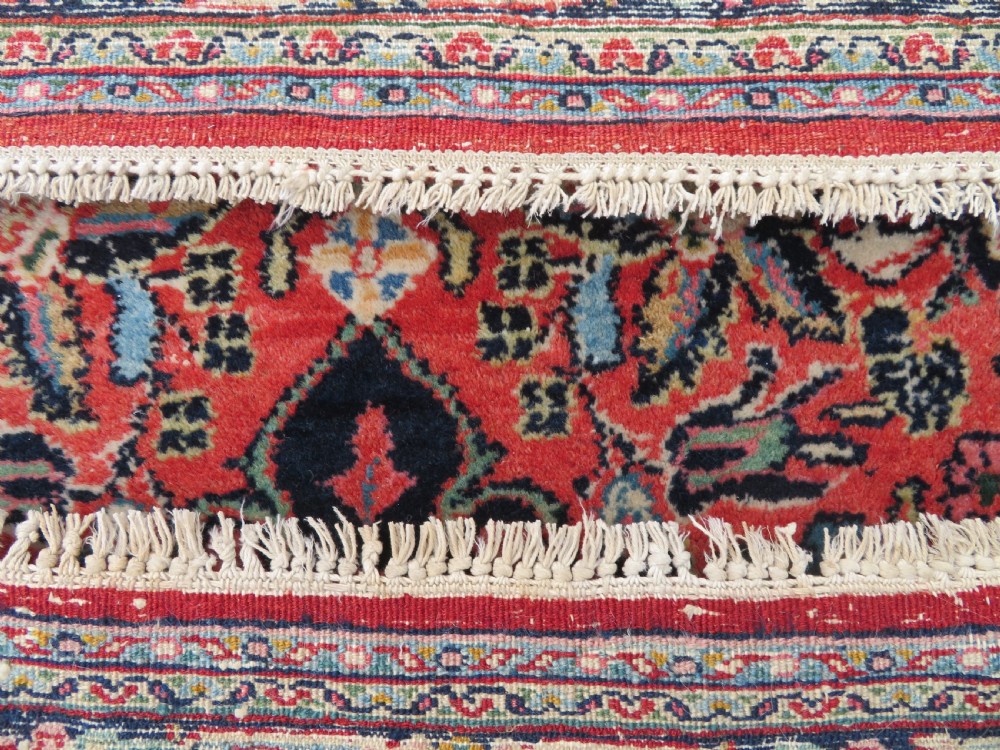 An antique hand-knotted Persian Sarouk wool rug featuring a red field with a central navy medallion and dense floral motifs within a dark border. View XV