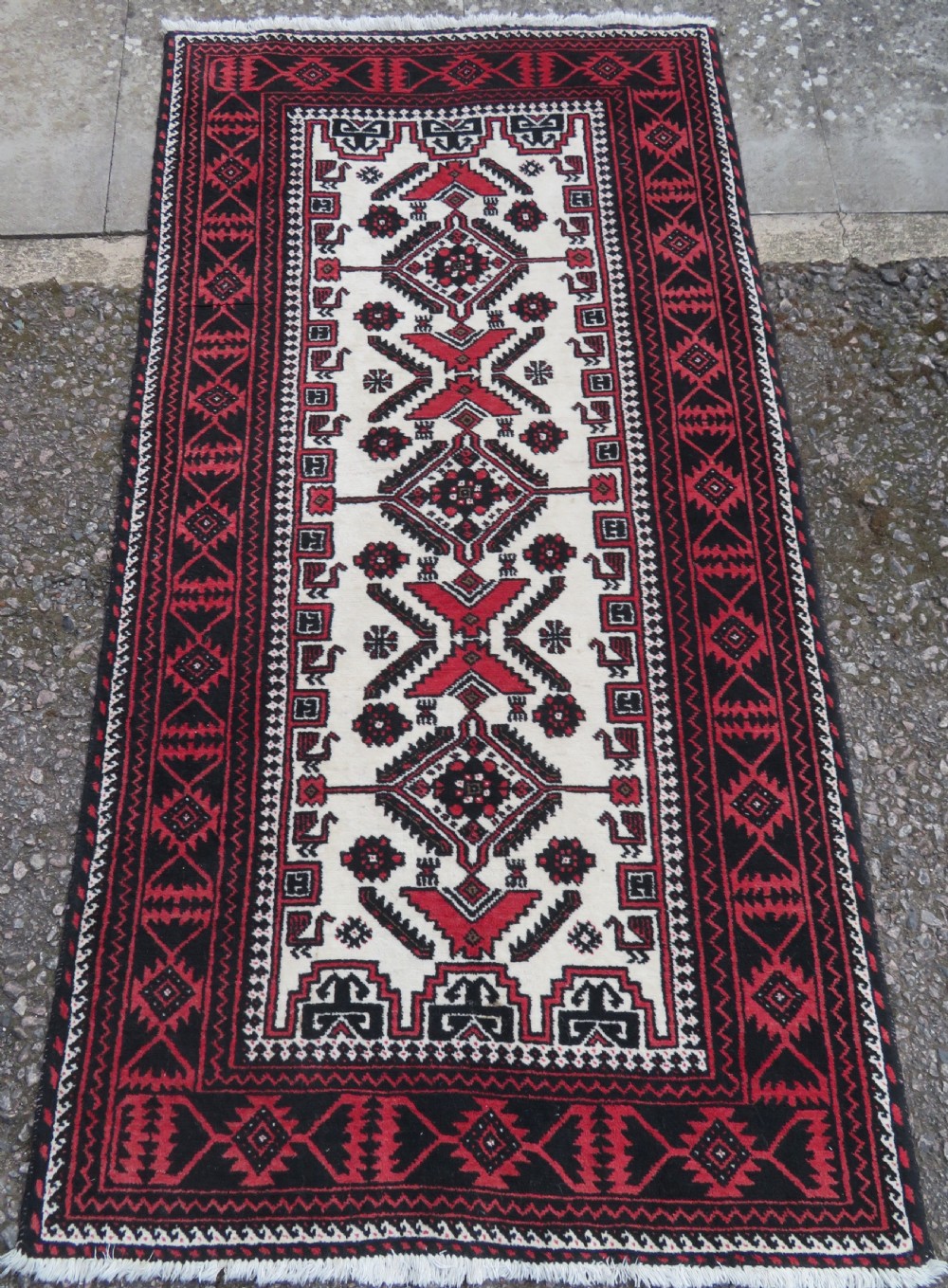 A rectangular hand-knotted wool Baluch rug featuring a central cream field with red geometric diamond motifs surrounded by a dark geometric border.