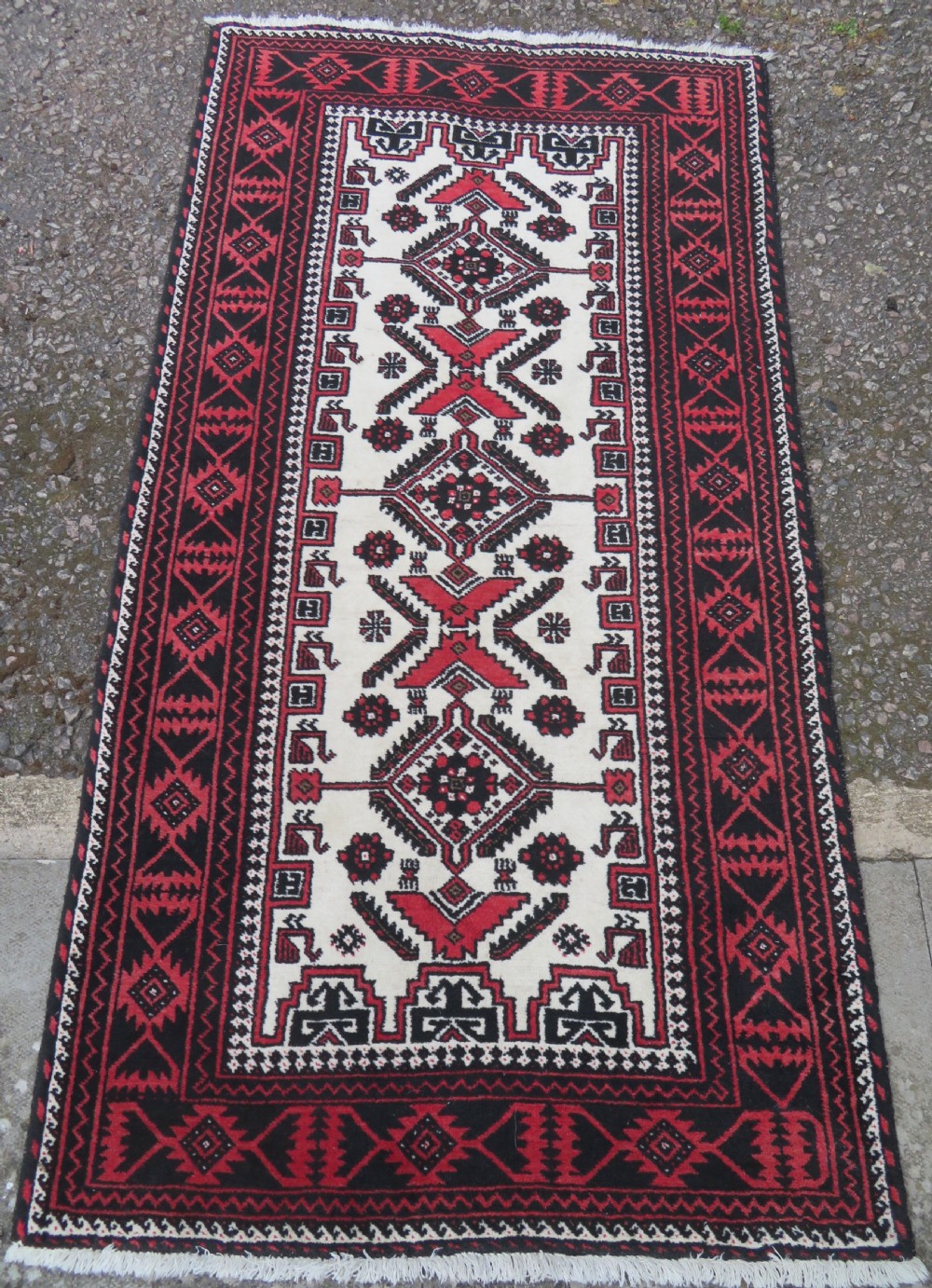 A rectangular hand-knotted wool Baluch rug featuring a central cream field with red geometric diamond motifs surrounded by a dark geometric border. View II