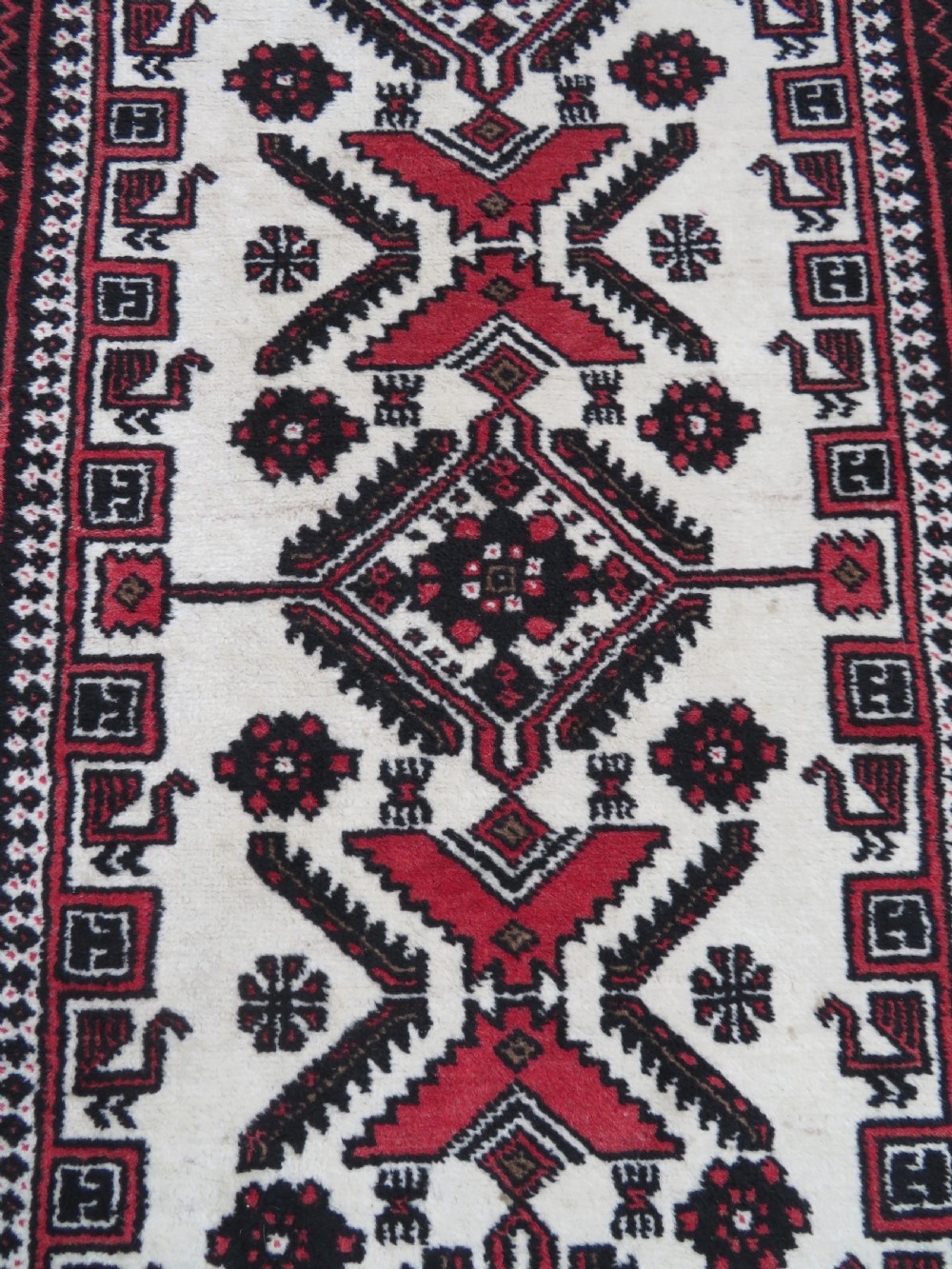 A rectangular hand-knotted wool Baluch rug featuring a central cream field with red geometric diamond motifs surrounded by a dark geometric border. View III