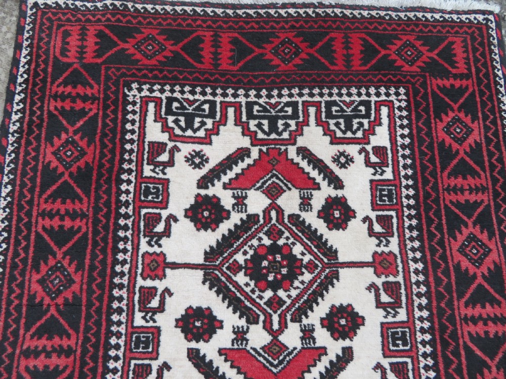 A rectangular hand-knotted wool Baluch rug featuring a central cream field with red geometric diamond motifs surrounded by a dark geometric border. View IV