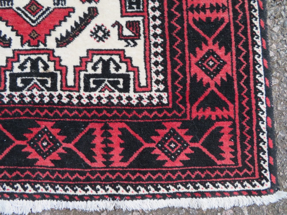 A rectangular hand-knotted wool Baluch rug featuring a central cream field with red geometric diamond motifs surrounded by a dark geometric border. View V