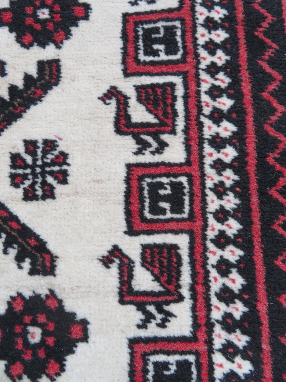 A rectangular hand-knotted wool Baluch rug featuring a central cream field with red geometric diamond motifs surrounded by a dark geometric border. View VI