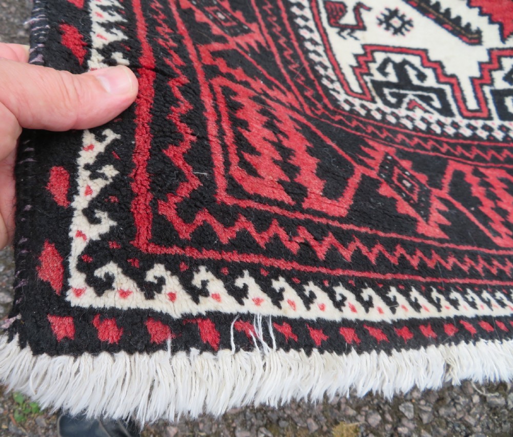 A rectangular hand-knotted wool Baluch rug featuring a central cream field with red geometric diamond motifs surrounded by a dark geometric border. View VII