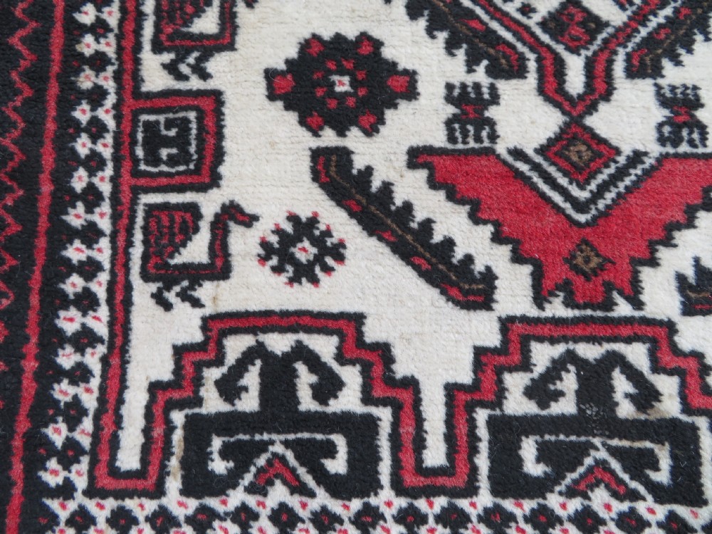 A rectangular hand-knotted wool Baluch rug featuring a central cream field with red geometric diamond motifs surrounded by a dark geometric border. View VIII