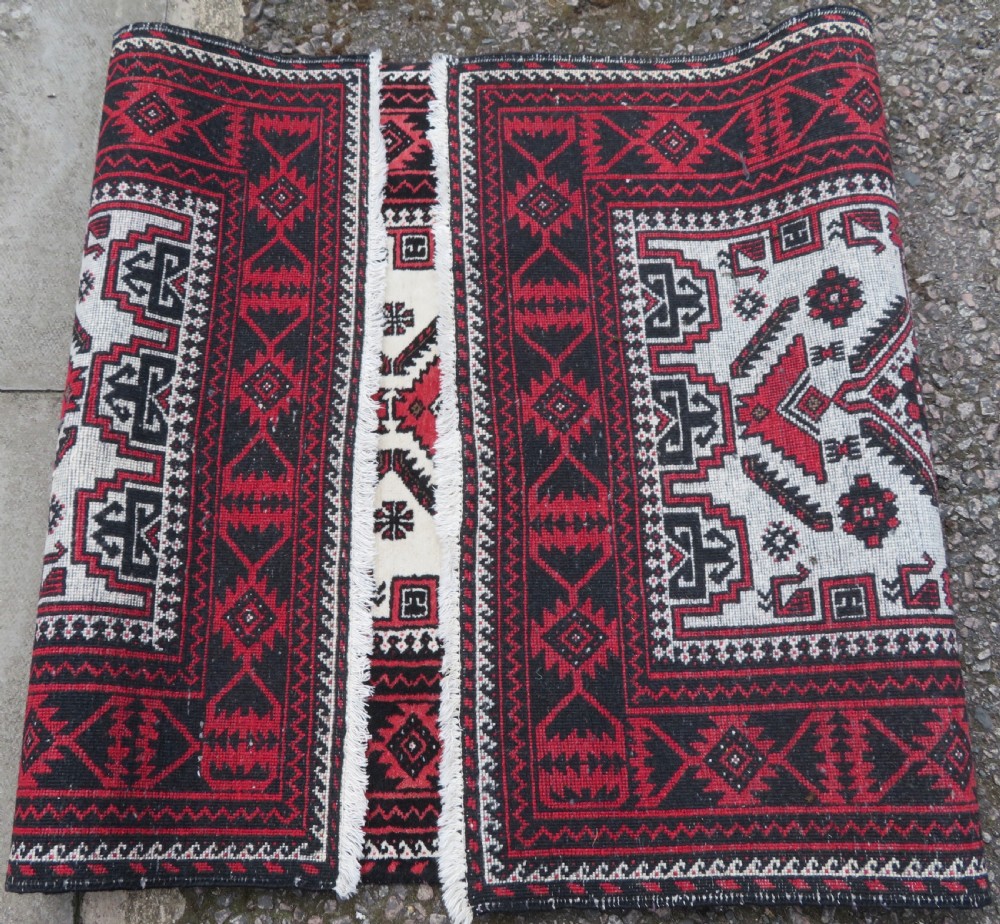 A rectangular hand-knotted wool Baluch rug featuring a central cream field with red geometric diamond motifs surrounded by a dark geometric border. View X