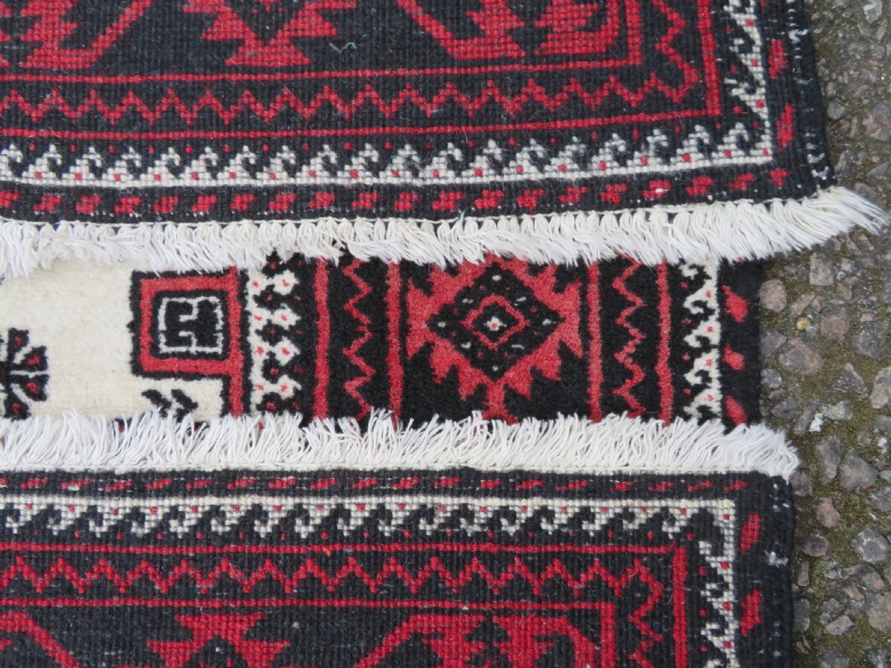 A rectangular hand-knotted wool Baluch rug featuring a central cream field with red geometric diamond motifs surrounded by a dark geometric border. View XI