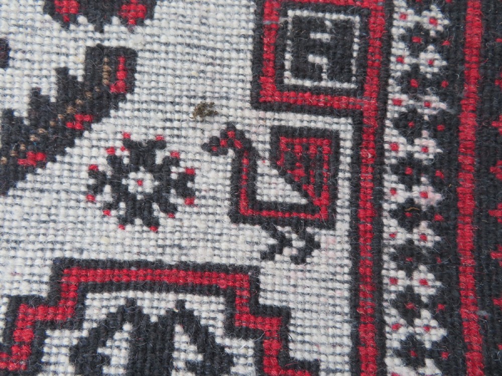 A rectangular hand-knotted wool Baluch rug featuring a central cream field with red geometric diamond motifs surrounded by a dark geometric border. View XII