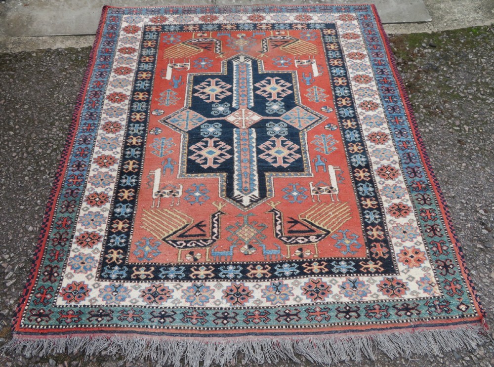 A handwoven rectangular wool rug featuring a central navy blue stepped medallion and a terracotta field decorated with peacock and animal motifs.